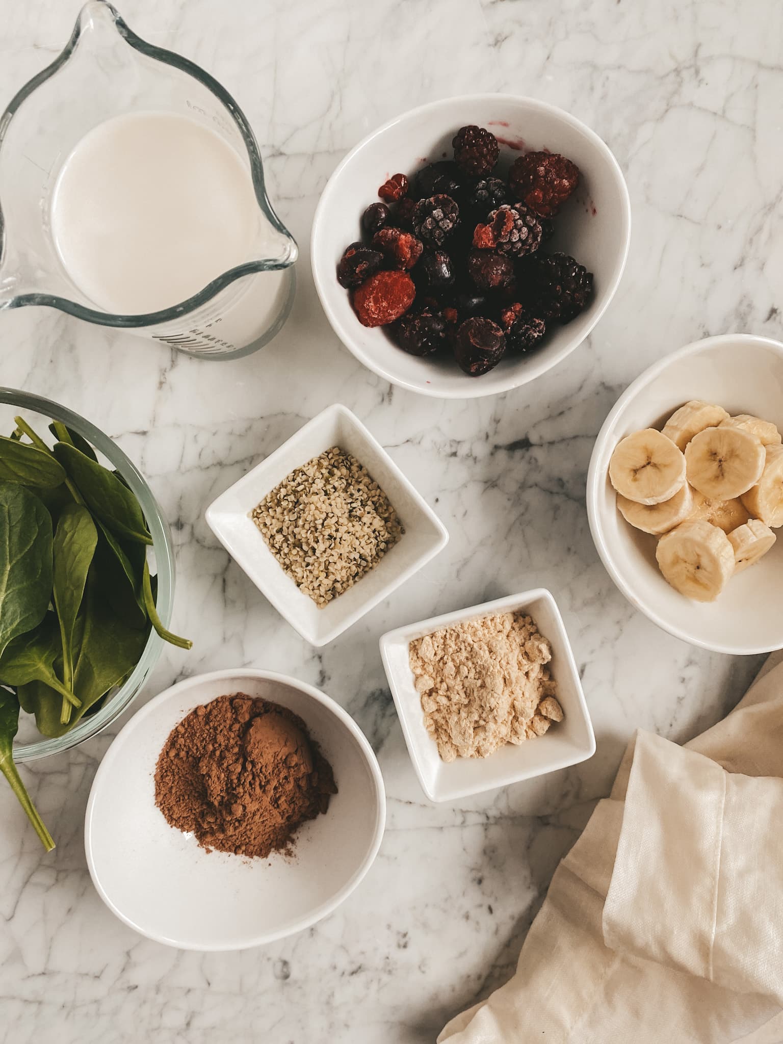 assorted bowls on a marble table with smoothie ingredients: spinach, almond milk, banana, cherries, hemp seeds, protein powder, and cocoa powder