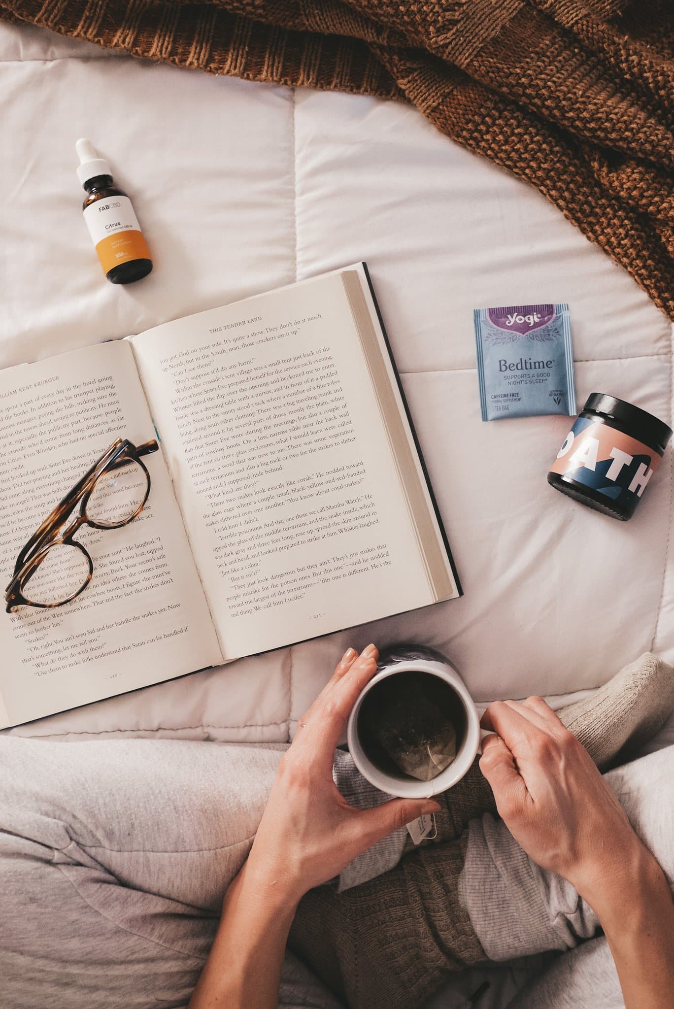 a white bedspread with a girl in gray sweatpants and taupe socks holding a mug of tea; a book, body lotion, package of tea, glasses and CBD bottle are on the bedspread