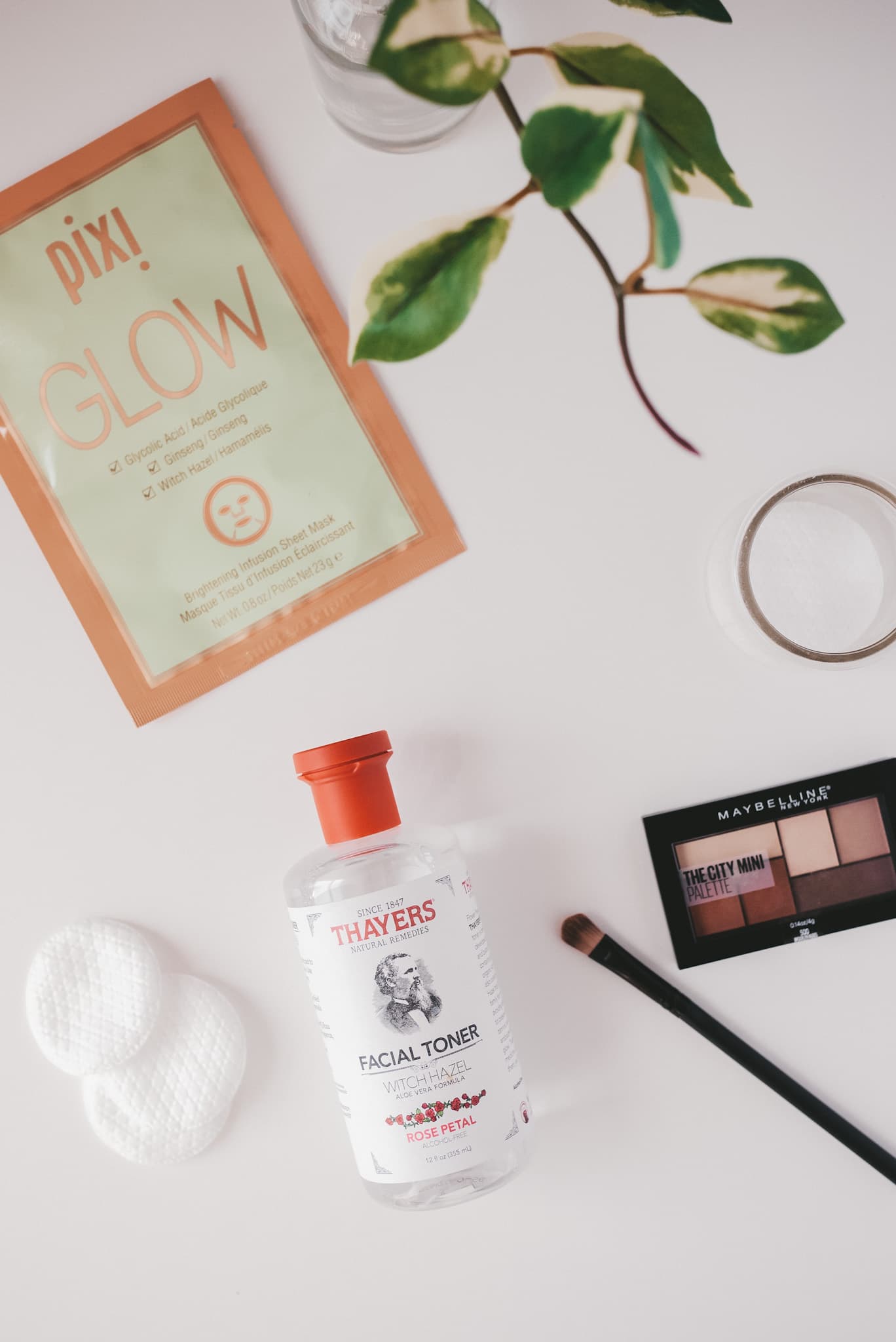 a white background with a Pixi Glow sheet mask, Thayer's Facial Toner, and Maybelline eye shadow palatte