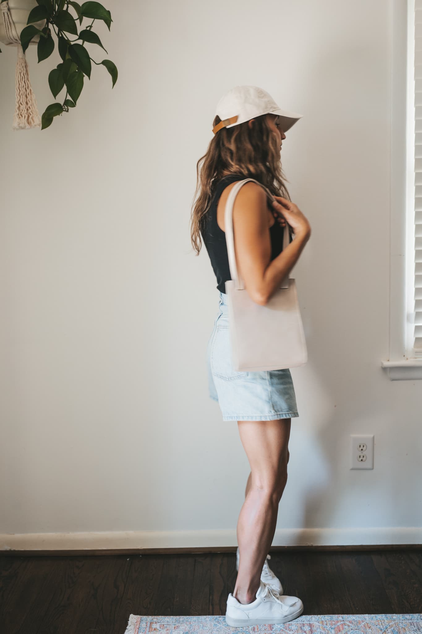 ashley wearing a black ribbed tank, light wash denim shorts, white sneakers, a beige baseball cap, and a gray leather shoulder bag
