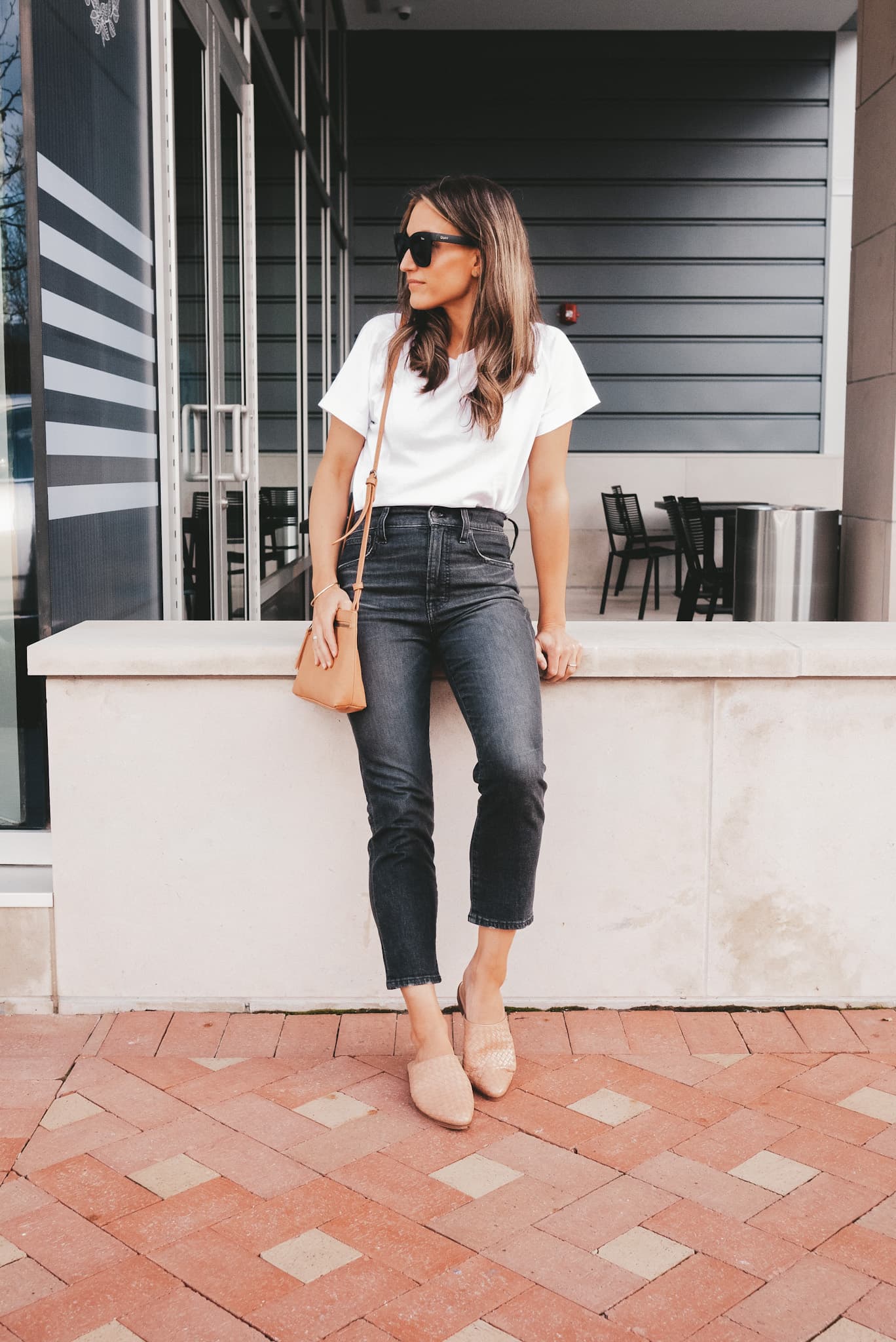 ashley wearing a white tee, washed black jeans and woven mules leaning against a concrete wall next to a coffee shop
