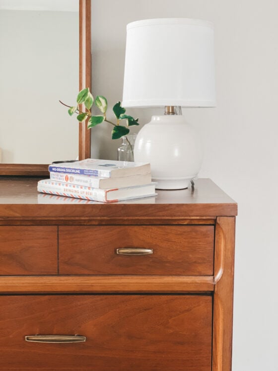 a wood dresser with a mirror, three books on the dresser with a small vase and green plant, and a white lamp