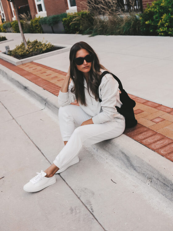 ashley sitting on a curb wearing a gray half zip sweatshirt, white joggers, and white sneakers