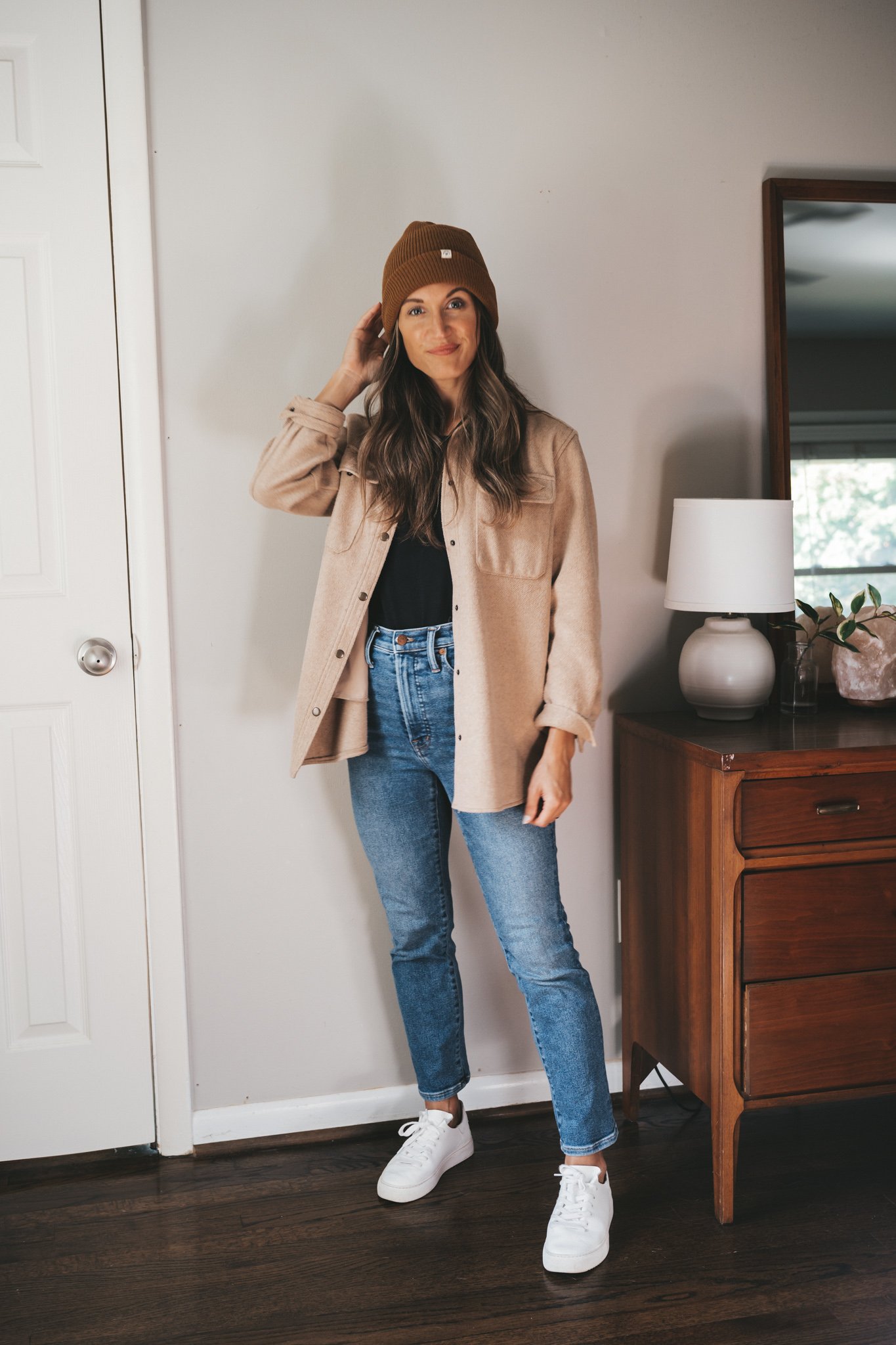 ashley wearing a black tank, tan shacket, medium wash denim, and white sneakers with a cedar colored beanie