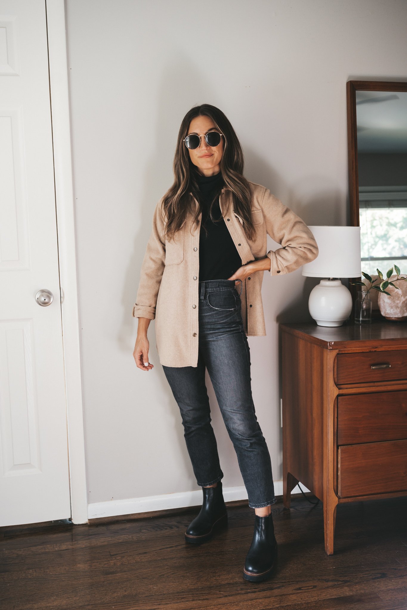 ashley wearing a black turtleneck, tan shacket, washed black jeans, black Chelsea boots, and round gold frame sunglasses