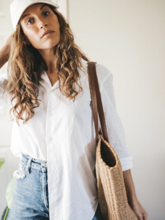 ashley wearing a white button down, denim shorts, white sandals, a woven tote bag, and a cream colored baseball cap.
