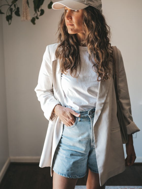 ashley wearing a white tee, beige oversized blazer, light wash long length denim shorts, white sneakers and a beige baseball cap with a gray shoulder bag