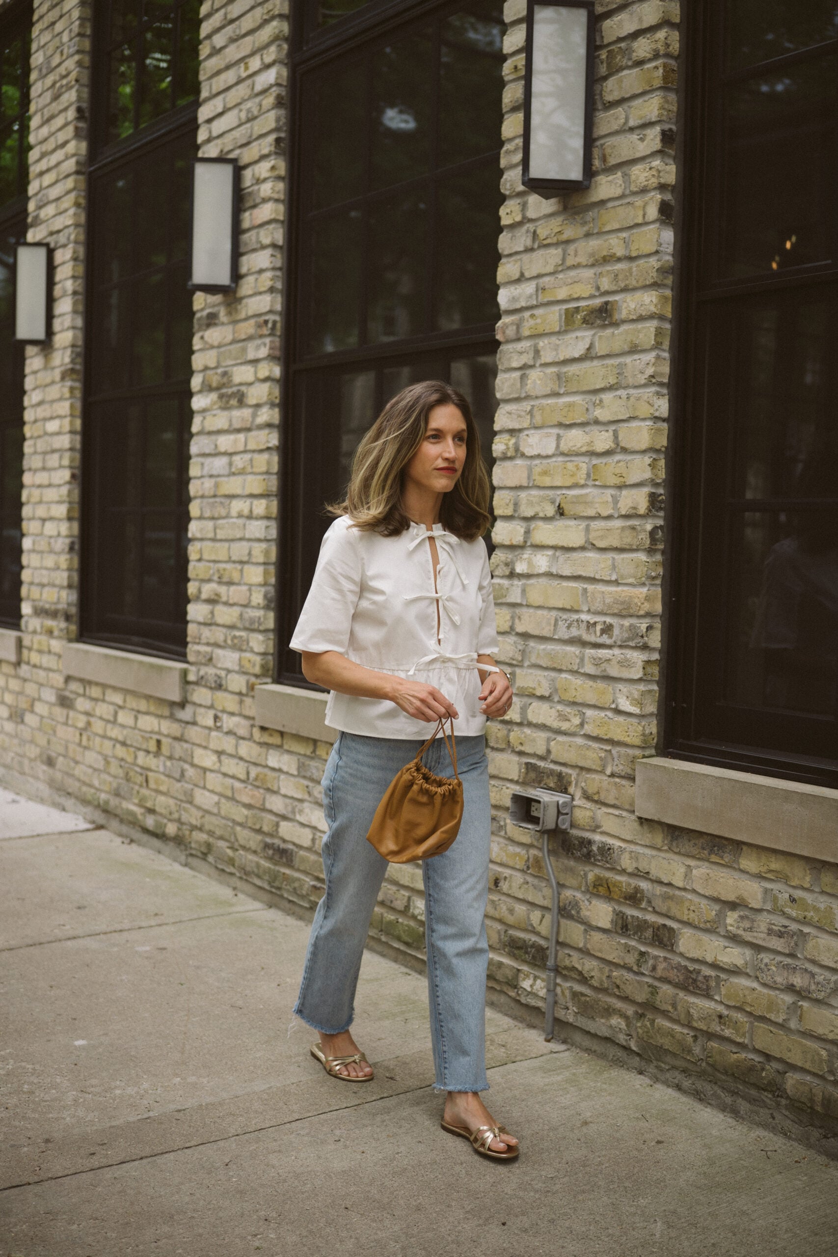 Quince rigid denim review - ashley wearing a white blouse with ties in the front, Quince rigid denim in a light wash, and gold sandals